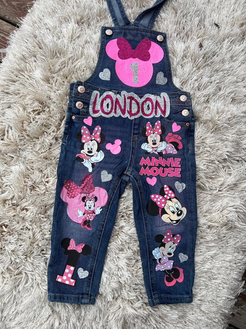 K&ouml;nnte beinhalten: Dunkelblaue Jeans-Latzhose mit Minnie-Maus-Motiven, rosa Schleifen und dem Namen "LONDON". Die Latzhose hat einen gro&szlig;en rosa Minnie-Maus-Kopf mit einer Schleife und der Zahl "1". Silberne Herz-Akzente sind ebenfalls vorhanden.