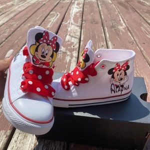 Puede incluir: Zapatillas blancas de ca&ntilde;a alta con cordones de cinta de lunares rojos y blancos. Las zapatillas tienen una imagen de dibujos animados de Minnie Mouse a cada lado. La zapatilla derecha tiene el nombre "Alayna" escrito en ella.