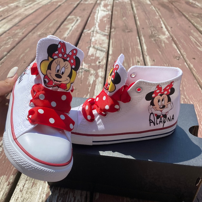 Puede incluir: Zapatillas blancas de ca&ntilde;a alta con cordones de cinta de lunares rojos y blancos. Las zapatillas tienen una imagen de dibujos animados de Minnie Mouse a cada lado. La zapatilla derecha tiene el nombre "Alayna" escrito en ella.