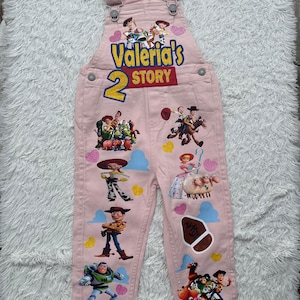 May include: Pale pink denim dungarees decorated with vibrant illustrations of characters from the animated film "Toy Story." The text "Valeria's Story" and the number "2" are printed on the front, alongside various character images.