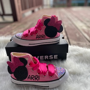 May include: Pink high-top Converse sneakers with a black Minnie Mouse ear applique and pink bow. The shoes are decorated with rhinestones and the name "ARRI" is written on the side.