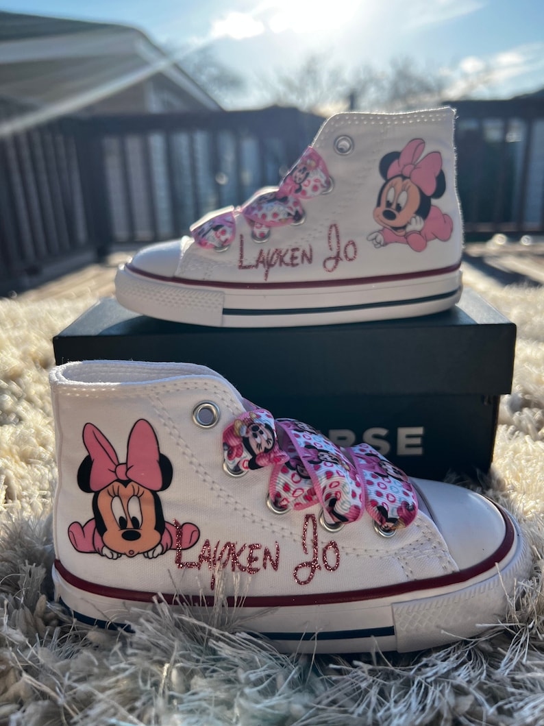 May include: White high-top sneakers with pink Minnie Mouse characters and pink polka dot ribbon laces. The shoes are personalized with the name "Layken Jo" in pink glitter.