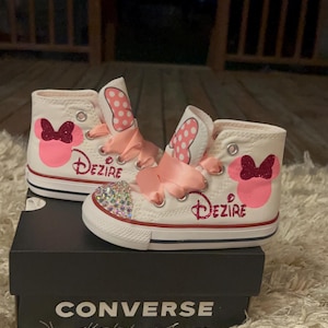 K&ouml;nnte beinhalten: Wei&szlig;e Converse High-Top-Sneaker mit einem Minnie-Maus-Design. Die Sneaker haben rosa Schleifen, den Namen "Dezire" in Glitzer und eine mit Strass besetzte Zehenkappe. Die Schuhe werden auf einer schwarzen Converse-Box pr&auml;sentiert.