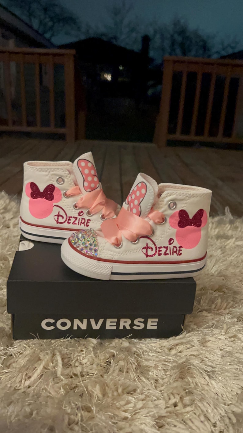 K&ouml;nnte beinhalten: Wei&szlig;e Converse High-Top-Sneaker mit einem Minnie-Maus-Design. Die Sneaker haben rosa Schleifen, den Namen "Dezire" in Glitzer und eine mit Strass besetzte Zehenkappe. Die Schuhe werden auf einer schwarzen Converse-Box pr&auml;sentiert.