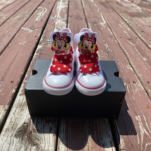 Puede incluir: Zapatillas blancas de ca&ntilde;a alta con cordones de cinta de lunares rojos y blancos y un personaje de Minnie Mouse en cada zapato.