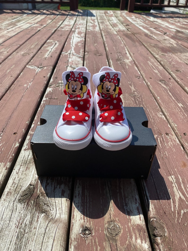 Puede incluir: Zapatillas blancas de ca&ntilde;a alta con cordones de cinta de lunares rojos y blancos y un personaje de Minnie Mouse en cada zapato.