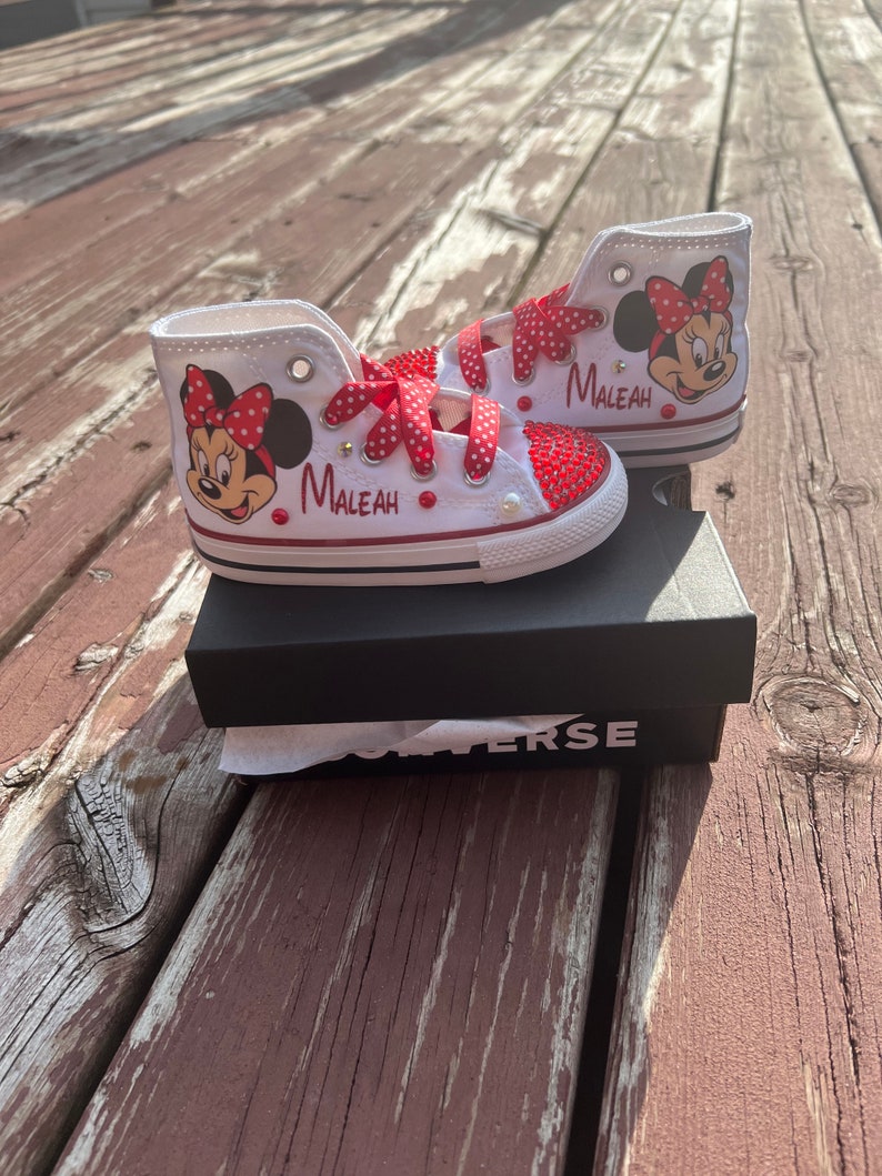 Peut inclure: Baskets montantes blanches avec un motif Minnie Mouse, des n&oelig;uds et des accents &agrave; pois rouges. Le nom "MALEAH" est imprim&eacute; sur le c&ocirc;t&eacute;. Les chaussures sont orn&eacute;es de strass rouges et de lacets &agrave; pois rouges. Les chaussures sont sur une bo&icirc;te noire.