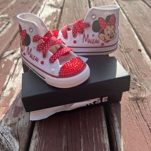 Peut inclure: Baskets montantes blanches avec des lacets rouges &agrave; pois et des motifs de Minnie Mouse. Les bouts des chaussures sont orn&eacute;s de strass rouges. Le nom "Maleah" est imprim&eacute; sur le c&ocirc;t&eacute; de chaque chaussure. Les chaussures sont pr&eacute;sent&eacute;es sur une bo&icirc;te Converse noire.