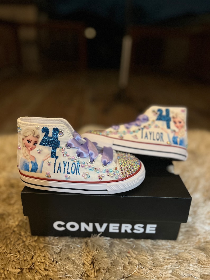 Puede incluir: Zapatillas Converse blancas de ca&ntilde;a alta con una imagen azul y blanca de Elsa de Frozen, un n&uacute;mero "4" de purpurina azul y el nombre "Taylor" en purpurina azul. Las zapatillas est&aacute;n sobre una caja negra con la palabra "CONVERSE" impresa en blanco.