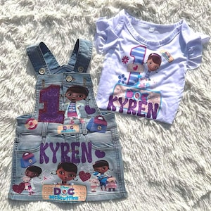May include: A denim overall dress and a white short-sleeve shirt featuring a cartoon character design. The overall dress has a purple glitter number "1" and the name "Kyren". The shirt also has the number "1" and the name "Kyren".
