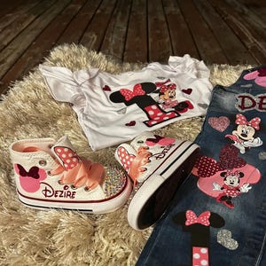 K&ouml;nnte beinhalten: Ein Set Kinderkleidung mit Minnie Maus-Motiv. Enth&auml;lt ein wei&szlig;es T-Shirt, Jeans und High-Top-Sneaker. Die Sneaker haben rosa Schleifen und den Namen "Dezire". Die Jeans und das Shirt haben Minnie Maus-Grafiken.