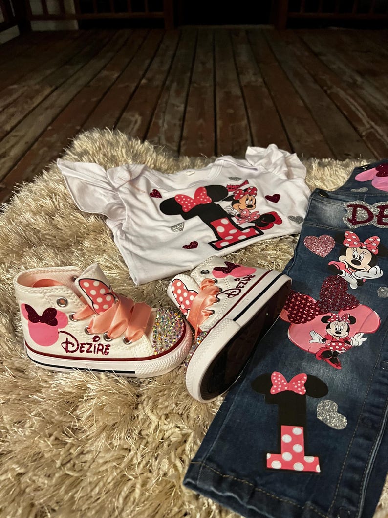 K&ouml;nnte beinhalten: Ein Set Kinderkleidung mit Minnie Maus-Motiv. Enth&auml;lt ein wei&szlig;es T-Shirt, Jeans und High-Top-Sneaker. Die Sneaker haben rosa Schleifen und den Namen "Dezire". Die Jeans und das Shirt haben Minnie Maus-Grafiken.