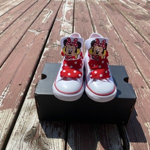 Puede incluir: Zapatillas blancas de ca&ntilde;a alta con lazos de lunares rojos y blancos y un gr&aacute;fico de Minnie Mouse en cada zapato.