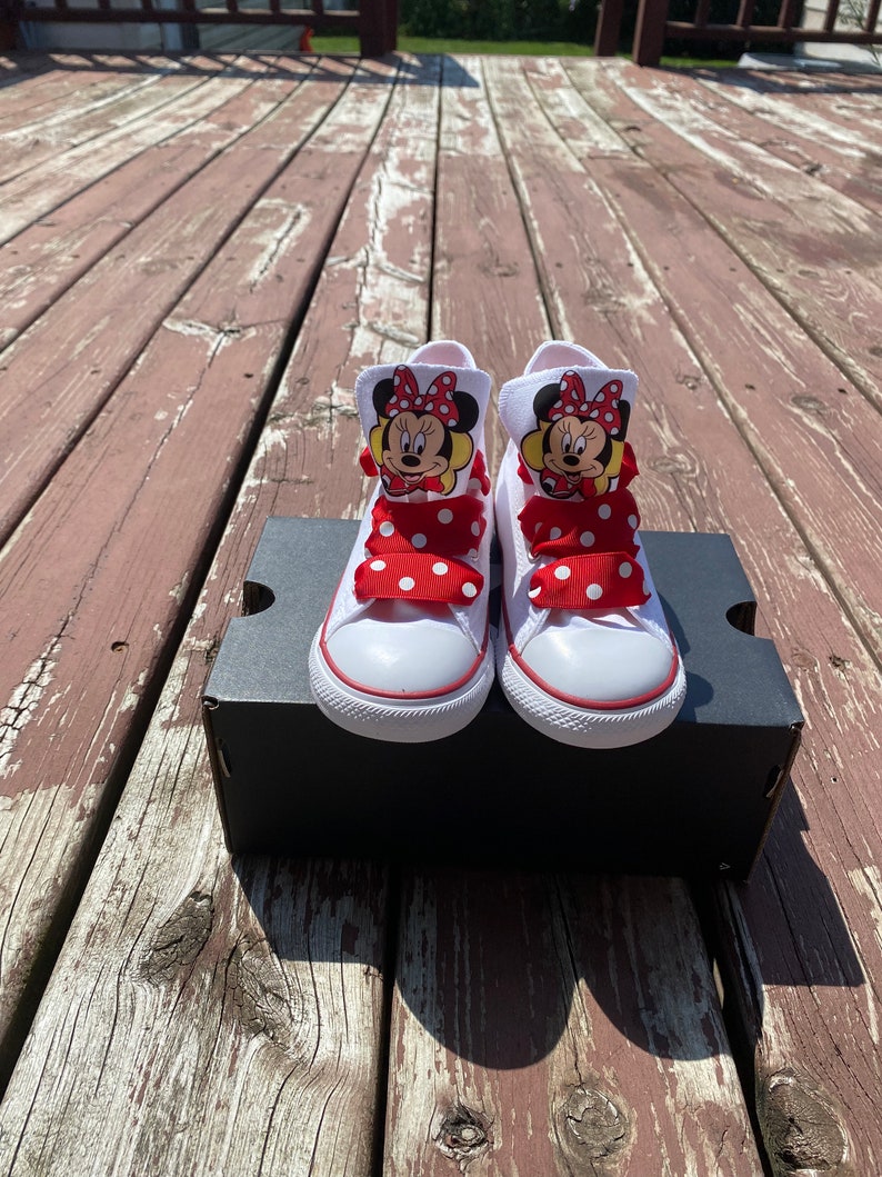Puede incluir: Zapatillas blancas de ca&ntilde;a alta con lazos de lunares rojos y blancos y un gr&aacute;fico de Minnie Mouse en cada zapato.