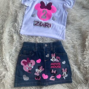 Puede incluir: Una camiseta blanca con una cabeza de Minnie Mouse rosa y el n&uacute;mero 2 en purpurina plateada. La camiseta dice "Zuri" en purpurina plateada. Una falda vaquera con aplicaciones de Minnie Mouse y el n&uacute;mero 2 en purpurina rosa. La falda dice "Minnie Mouse" en negro.