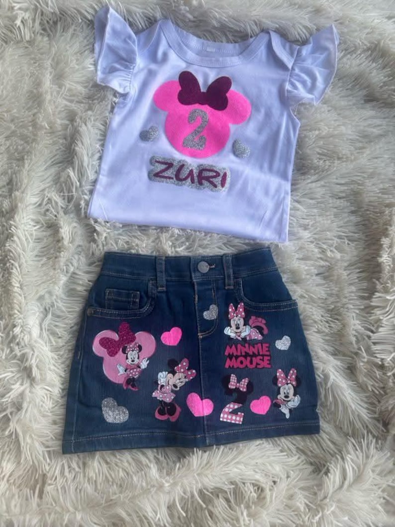 Puede incluir: Una camiseta blanca con una cabeza de Minnie Mouse rosa y el n&uacute;mero 2 en purpurina plateada. La camiseta dice "Zuri" en purpurina plateada. Una falda vaquera con aplicaciones de Minnie Mouse y el n&uacute;mero 2 en purpurina rosa. La falda dice "Minnie Mouse" en negro.