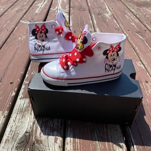 Puede incluir: Zapatillas blancas de ca&ntilde;a alta con lazos de lunares rojos y blancos. Las zapatillas tienen un gr&aacute;fico de Minnie Mouse a cada lado y el nombre "ALAYNA" escrito en el lateral.