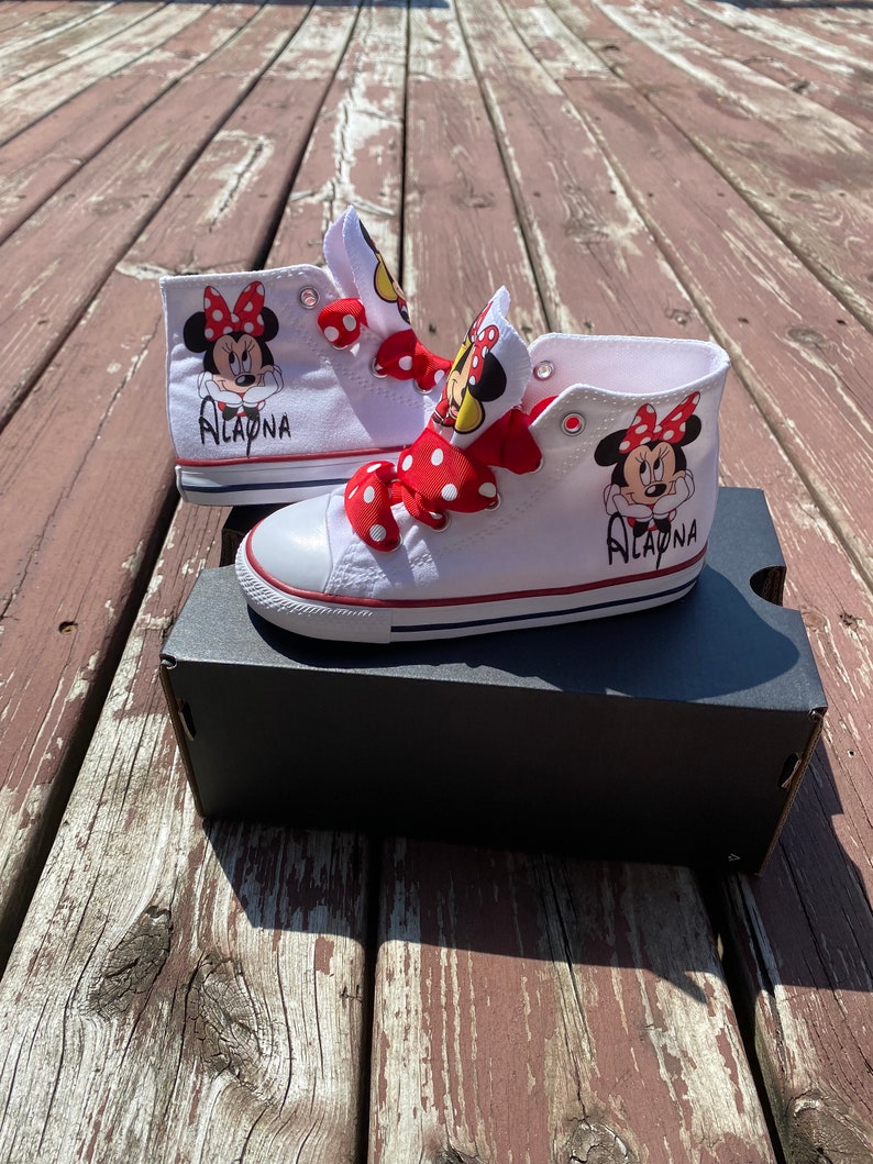 Puede incluir: Zapatillas blancas de ca&ntilde;a alta con lazos de lunares rojos y blancos. Las zapatillas tienen un gr&aacute;fico de Minnie Mouse a cada lado y el nombre "ALAYNA" escrito en el lateral.