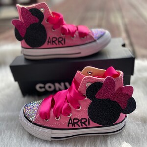 May include: Pink high-top Converse sneakers with a sparkly pink bow and black Mickey Mouse ears. The shoes are personalized with the name "ARRI" in black lettering.
