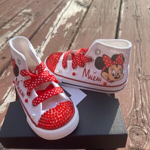Peut inclure: Baskets montantes blanches avec des accents de strass rouges et des lacets &agrave; pois rouges. Comprend un dessin de Minnie Mouse et le nom "MALEAH" en noir. Les chaussures sont sur une bo&icirc;te noire.