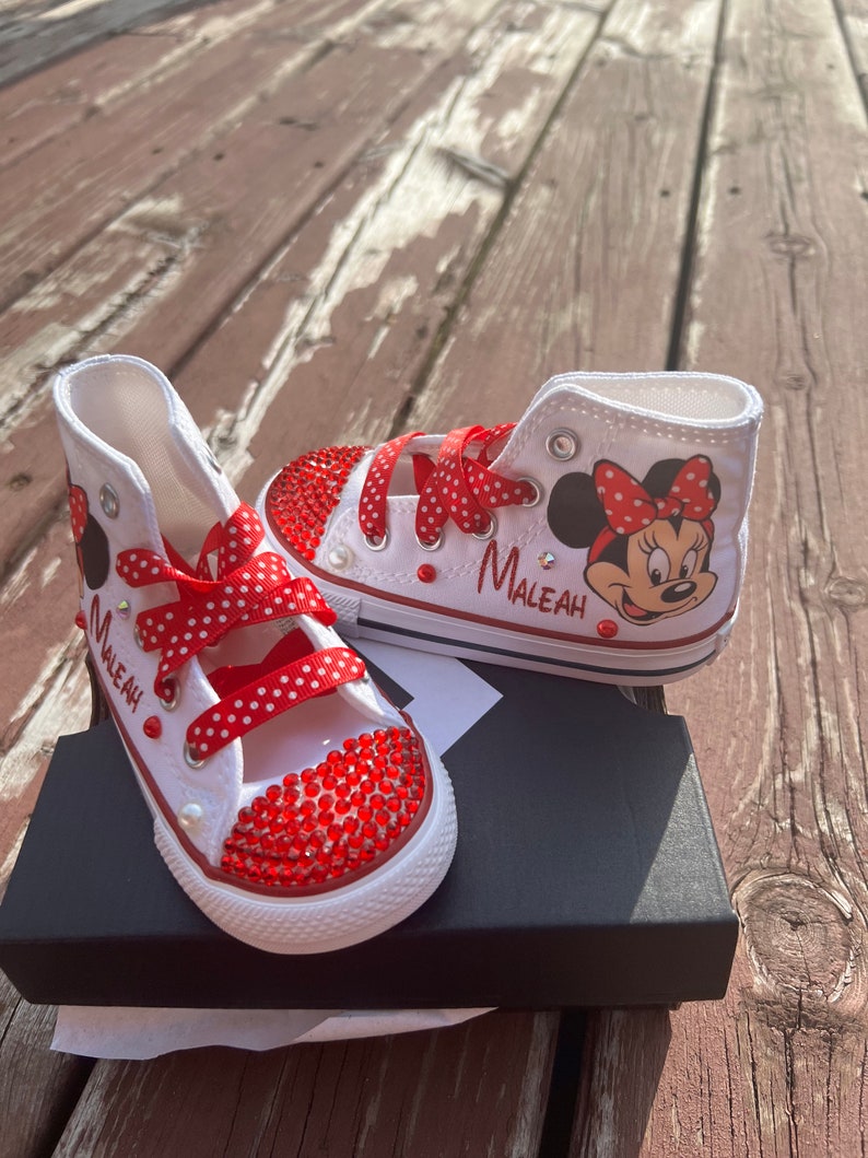 Peut inclure: Baskets montantes blanches avec des accents de strass rouges et des lacets &agrave; pois rouges. Comprend un dessin de Minnie Mouse et le nom "MALEAH" en noir. Les chaussures sont sur une bo&icirc;te noire.