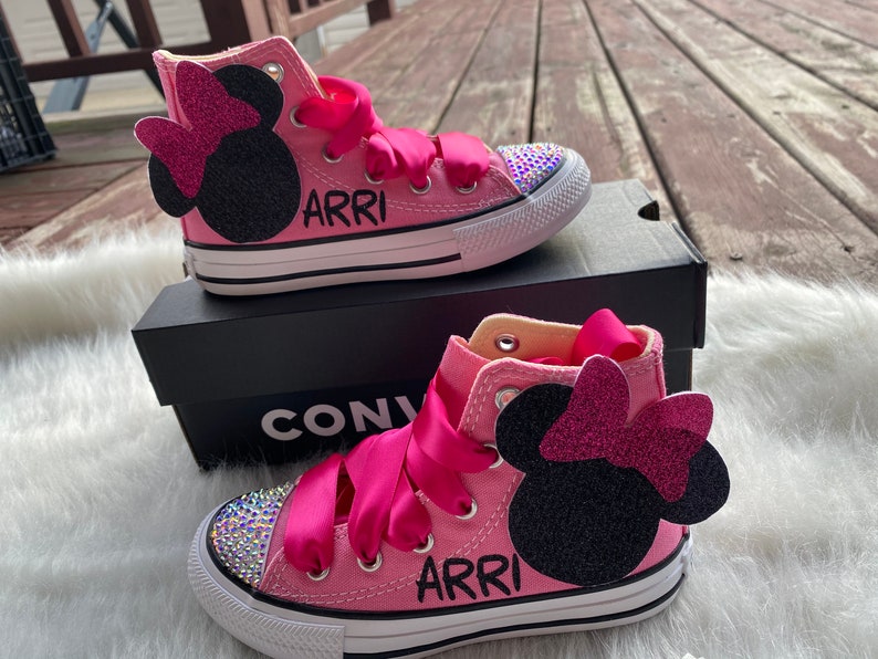 May include: Pink high-top Converse sneakers with a black Minnie Mouse design and pink bow. The shoes have a white sole and are decorated with rhinestones. The name "ARRI" is written on the side of the shoes.