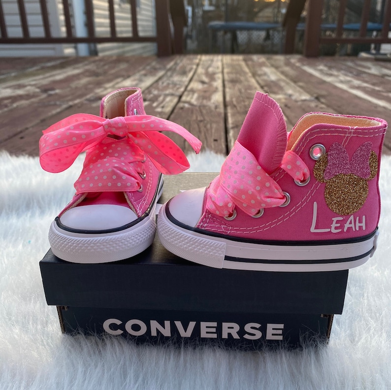 Puede incluir: Zapatillas Converse de ca&ntilde;a alta de color rosa con un lazo blanco de lunares, una cabeza de Minnie Mouse de purpurina dorada con un lazo rosa y el nombre "Leah" escrito en el lateral. Las zapatillas est&aacute;n sobre una caja Converse negra.
