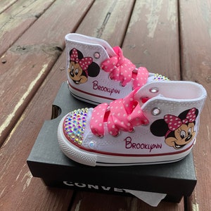 May include: White high-top Converse sneakers with pink polka dot ribbon laces and a Minnie Mouse design. The shoes are personalized with the name "Brooklynn" and have rhinestones on the toe.