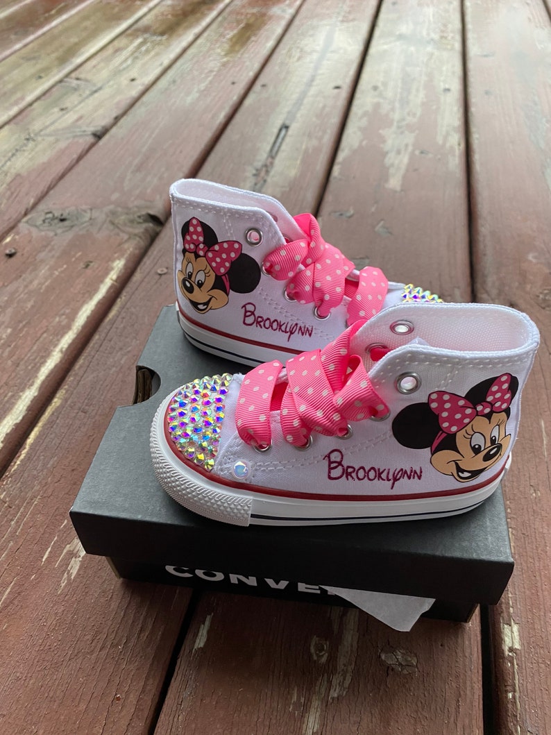 May include: White high-top Converse sneakers with pink polka dot ribbon laces and a Minnie Mouse design. The shoes are personalized with the name "Brooklynn" and have rhinestones on the toe.