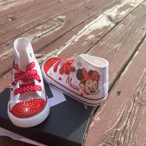 Peut inclure: Baskets montantes blanches avec des strass rouges et des lacets &agrave; pois rouges. Une chaussure pr&eacute;sente un dessin de Minnie Mouse et le nom "Maleah". Les chaussures sont sur une bo&icirc;te noire, sur un fond en bois.