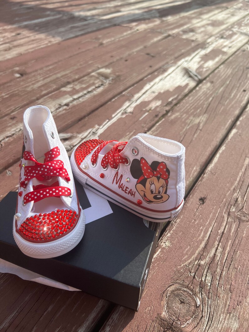 Peut inclure: Baskets montantes blanches avec des strass rouges et des lacets &agrave; pois rouges. Une chaussure pr&eacute;sente un dessin de Minnie Mouse et le nom "Maleah". Les chaussures sont sur une bo&icirc;te noire, sur un fond en bois.