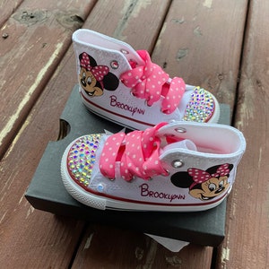 May include: White high-top sneakers with pink polka dot ribbon laces and a Minnie Mouse design. The shoes are decorated with rhinestones and the name "Brooklynn" is written on the side.