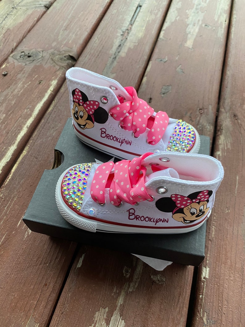 May include: White high-top sneakers with pink polka dot ribbon laces and a Minnie Mouse design. The shoes are decorated with rhinestones and the name "Brooklynn" is written on the side.