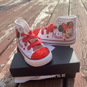 Peut inclure: Baskets montantes blanches avec des lacets rouges &agrave; pois et un bout rouge orn&eacute; de bijoux. Les baskets pr&eacute;sentent un dessin de Minnie Mouse et le nom "Maleah" en &eacute;criture rouge. Les chaussures sont pr&eacute;sent&eacute;es sur une bo&icirc;te noire.