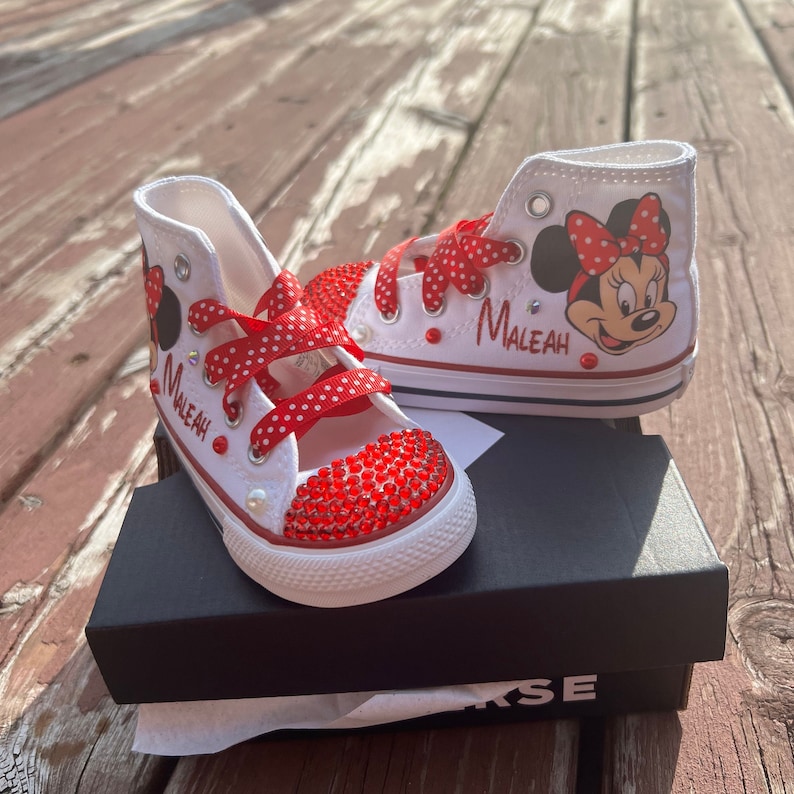 Peut inclure: Baskets montantes blanches avec des lacets rouges &agrave; pois et un bout rouge orn&eacute; de bijoux. Les baskets pr&eacute;sentent un dessin de Minnie Mouse et le nom "Maleah" en &eacute;criture rouge. Les chaussures sont pr&eacute;sent&eacute;es sur une bo&icirc;te noire.