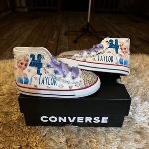 Puede incluir: Zapatillas Converse blancas de ca&ntilde;a alta con un dise&ntilde;o de personaje Frozen azul y blanco, pedrer&iacute;a y el nombre "Taylor" en el lateral. Las zapatillas est&aacute;n sobre una caja Converse negra.