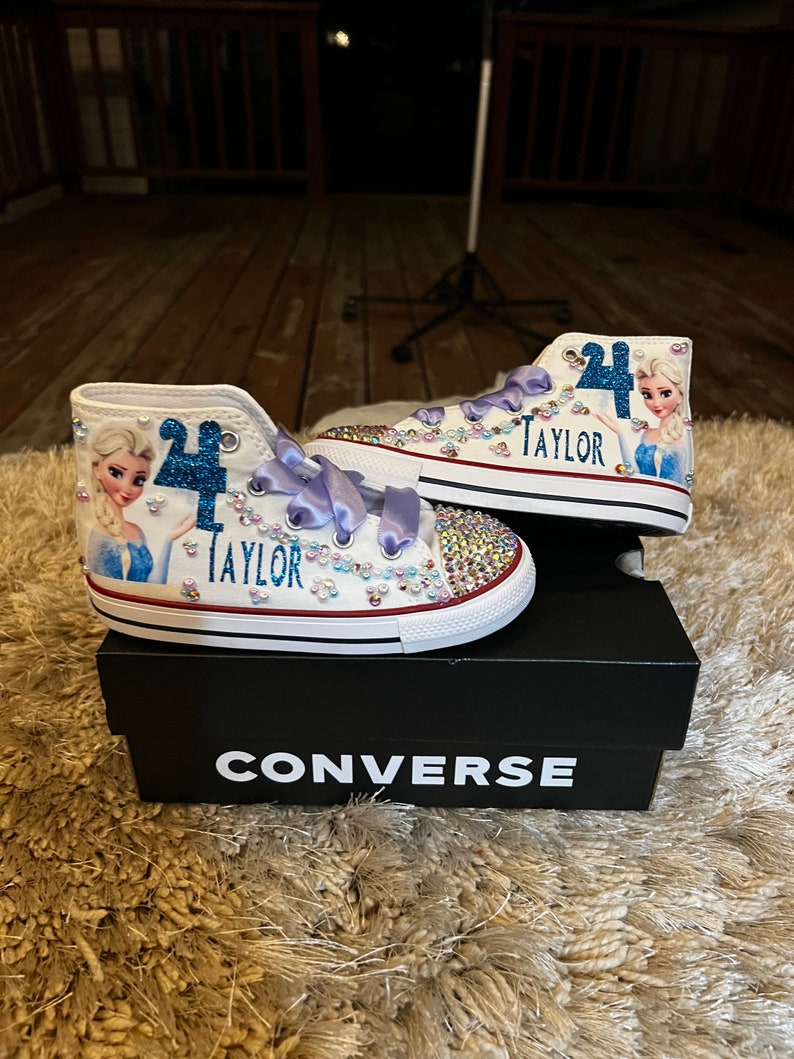 Puede incluir: Zapatillas Converse blancas de ca&ntilde;a alta con un dise&ntilde;o de personaje Frozen azul y blanco, pedrer&iacute;a y el nombre "Taylor" en el lateral. Las zapatillas est&aacute;n sobre una caja Converse negra.
