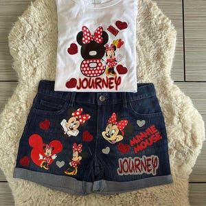 Peut inclure: Ensemble compos&eacute; d'un d&eacute;bardeur blanc et d'un short en jean avec des motifs de Minnie Mouse. Le d&eacute;bardeur pr&eacute;sente un grand motif de Minnie Mouse avec le mot "Journey". Le short en jean comporte plusieurs motifs de Minnie Mouse et les mots "Minnie Mouse Journey".