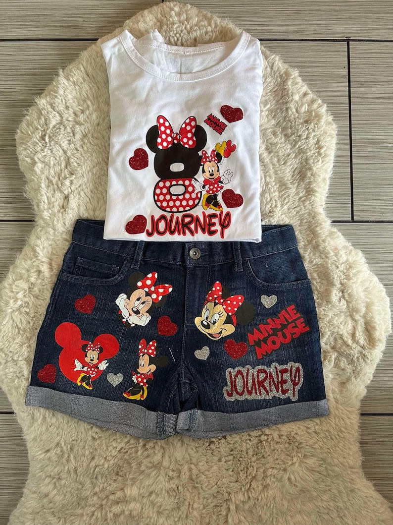 Peut inclure: Ensemble compos&eacute; d'un d&eacute;bardeur blanc et d'un short en jean avec des motifs de Minnie Mouse. Le d&eacute;bardeur pr&eacute;sente un grand motif de Minnie Mouse avec le mot "Journey". Le short en jean comporte plusieurs motifs de Minnie Mouse et les mots "Minnie Mouse Journey".
