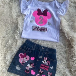 Puede incluir: Una camiseta blanca con una cabeza de Minnie Mouse rosa con un lazo burdeos y el n&uacute;mero 2 en purpurina plateada. La camiseta dice "Zuri" en purpurina plateada. Una falda vaquera con aplicaciones de Minnie Mouse y las palabras "Minnie Mouse" en blanco.
