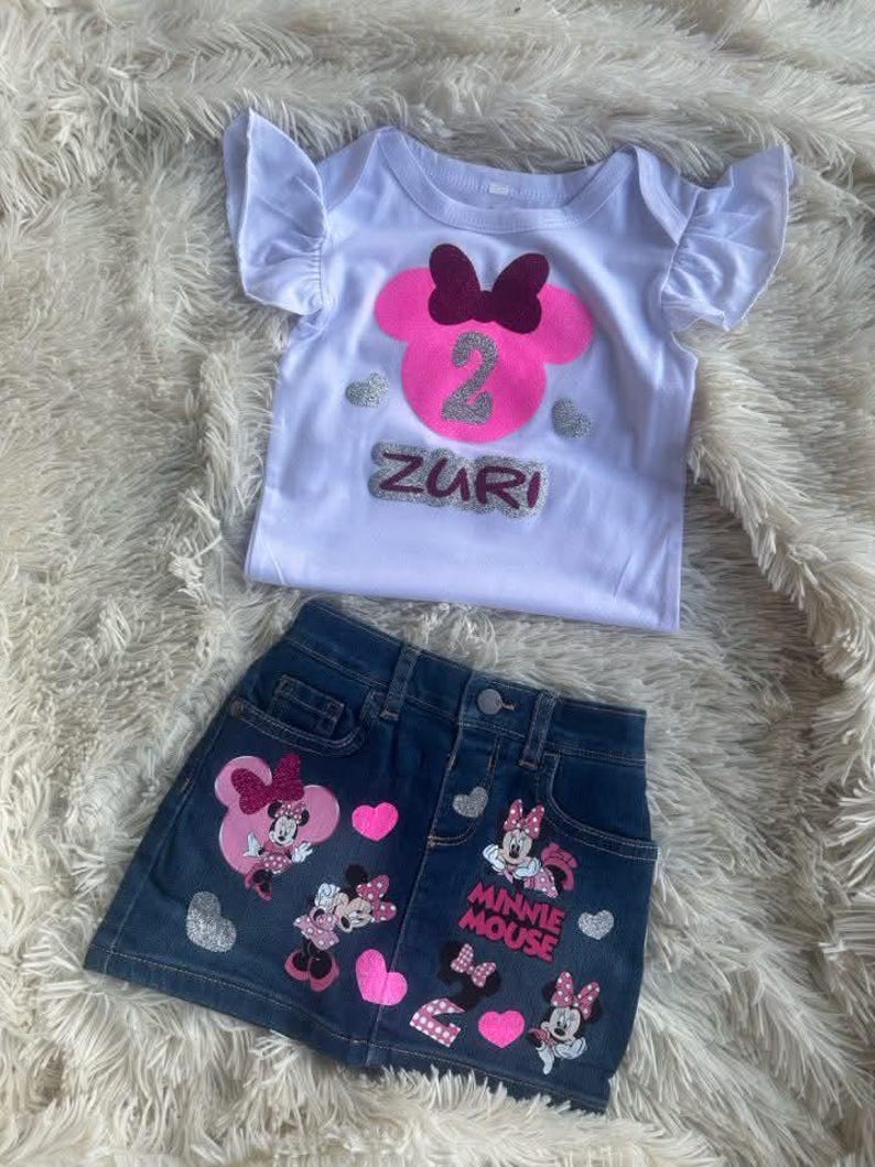 Puede incluir: Una camiseta blanca con una cabeza de Minnie Mouse rosa con un lazo burdeos y el n&uacute;mero 2 en purpurina plateada. La camiseta dice "Zuri" en purpurina plateada. Una falda vaquera con aplicaciones de Minnie Mouse y las palabras "Minnie Mouse" en blanco.