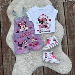 May include: A white t-shirt with Minnie Mouse and the text Oh Twodles, a pair of pink and blue denim overalls with Minnie Mouse graphics and the name Journee, and two white high-top sneakers with pink laces, Minnie Mouse, and the name Journee.