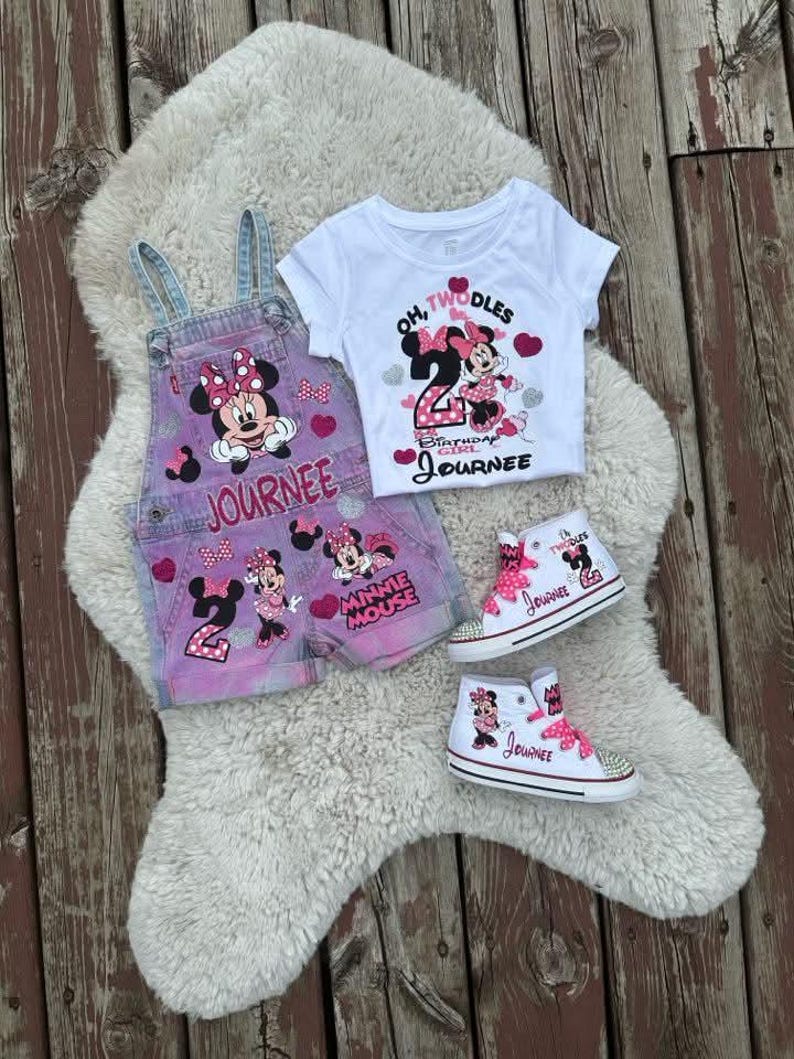 May include: A white t-shirt with Minnie Mouse and the text Oh Twodles, a pair of pink and blue denim overalls with Minnie Mouse graphics and the name Journee, and two white high-top sneakers with pink laces, Minnie Mouse, and the name Journee.