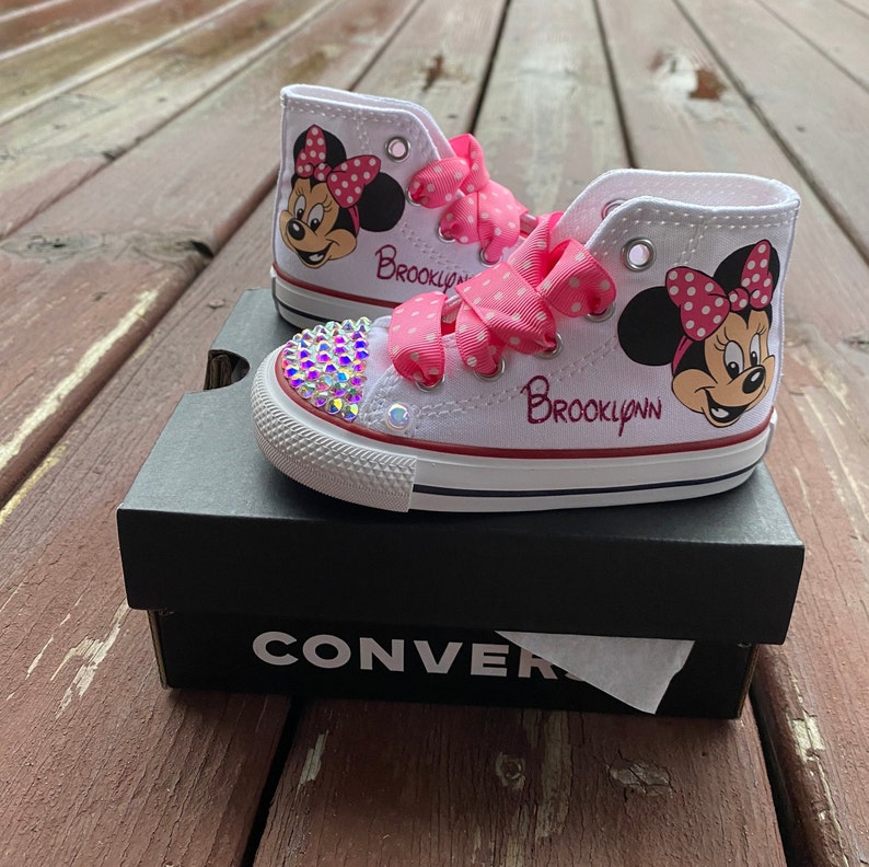 May include: White high-top Converse sneakers with a pink bow and Minnie Mouse design. The shoes are decorated with rhinestones and the name "Brooklynn" is written on the side.