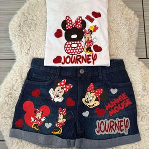 Peut inclure: Ensemble compos&eacute; d'un d&eacute;bardeur blanc et d'un short en jean avec des motifs de Minnie Mouse. Le haut pr&eacute;sente un grand motif de Minnie Mouse et le mot "Journey". Le short a plusieurs motifs de Minnie Mouse et les mots "Minnie Mouse Journey".
