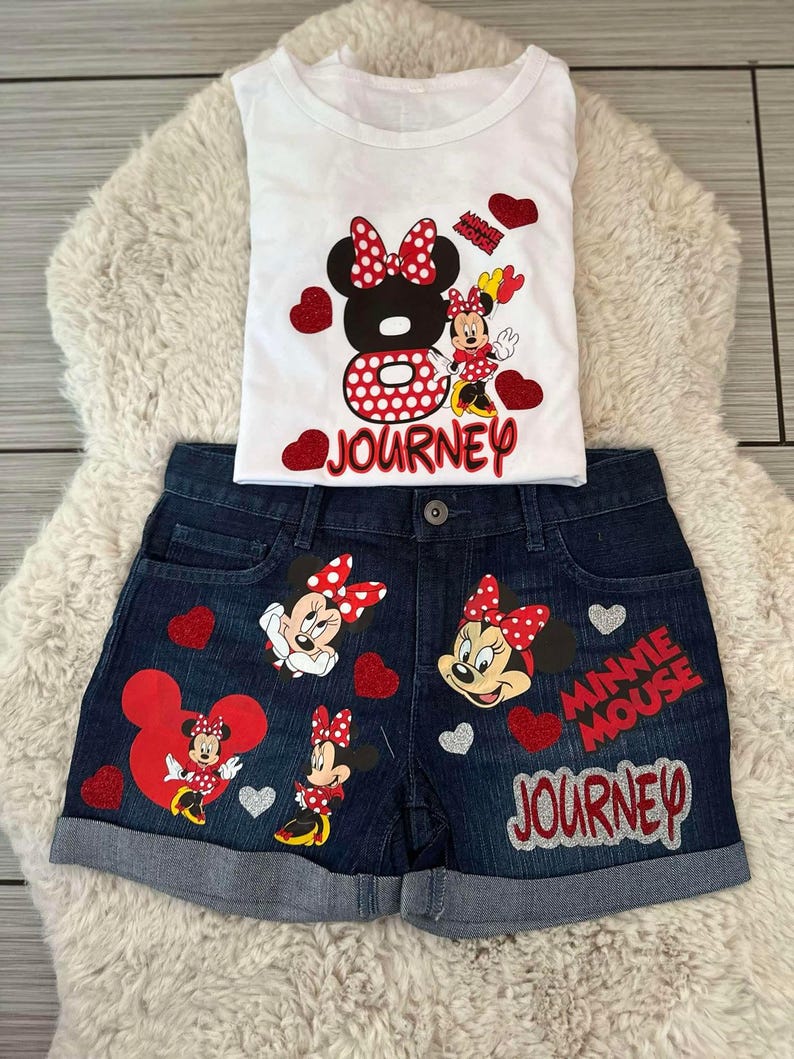Peut inclure: Ensemble compos&eacute; d'un d&eacute;bardeur blanc et d'un short en jean avec des motifs de Minnie Mouse. Le haut pr&eacute;sente un grand motif de Minnie Mouse et le mot "Journey". Le short a plusieurs motifs de Minnie Mouse et les mots "Minnie Mouse Journey".