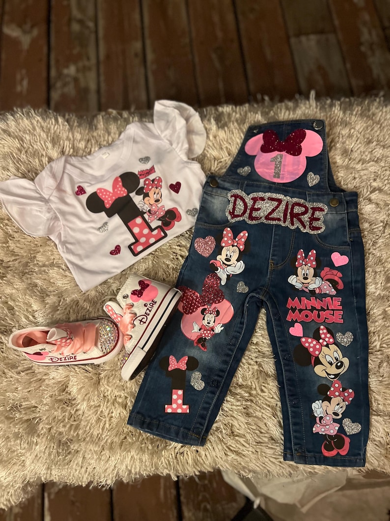K&ouml;nnte beinhalten: Ein Outfit mit Minnie Mouse-Design, bestehend aus einem wei&szlig;en T-Shirt, einer Jeans-Latzhose und rosa Schuhen. Das Outfit enth&auml;lt die Zahl "1" und den Namen "Dezire". Die High-Top-Sneakers und die Latzhose sind mit Glitzer verziert.