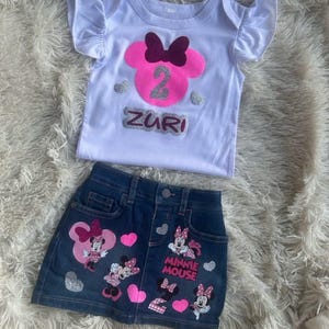 Puede incluir: Una camiseta blanca con una cabeza de Minnie Mouse rosa con un lazo burdeos y un n&uacute;mero 2 de purpurina plateada. La camiseta dice "Zuri" en letras de purpurina plateada. Una falda vaquera con corazones rosas y aplicaciones de Minnie Mouse. La falda dice "Minnie Mouse" en letras de purpurina plateada.