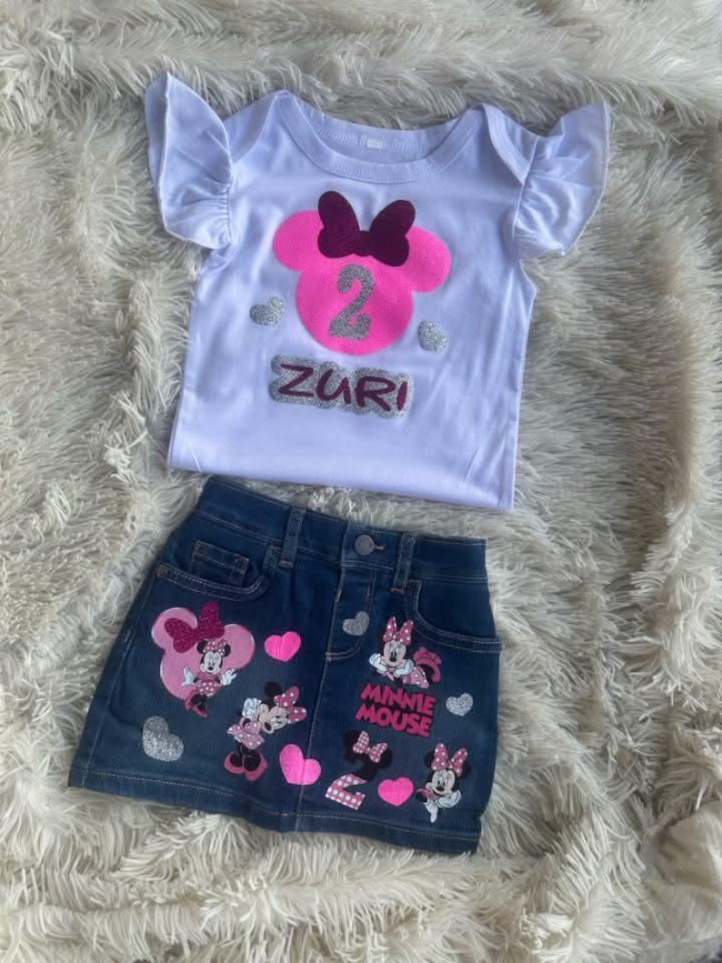 Puede incluir: Una camiseta blanca con una cabeza de Minnie Mouse rosa con un lazo burdeos y un n&uacute;mero 2 de purpurina plateada. La camiseta dice "Zuri" en letras de purpurina plateada. Una falda vaquera con corazones rosas y aplicaciones de Minnie Mouse. La falda dice "Minnie Mouse" en letras de purpurina plateada.