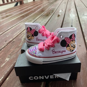 May include: White high-top sneakers with a pink polka dot ribbon, a Minnie Mouse design, and the name "Brooklyn" written on the side. The sneakers are sitting on a black Converse shoebox.